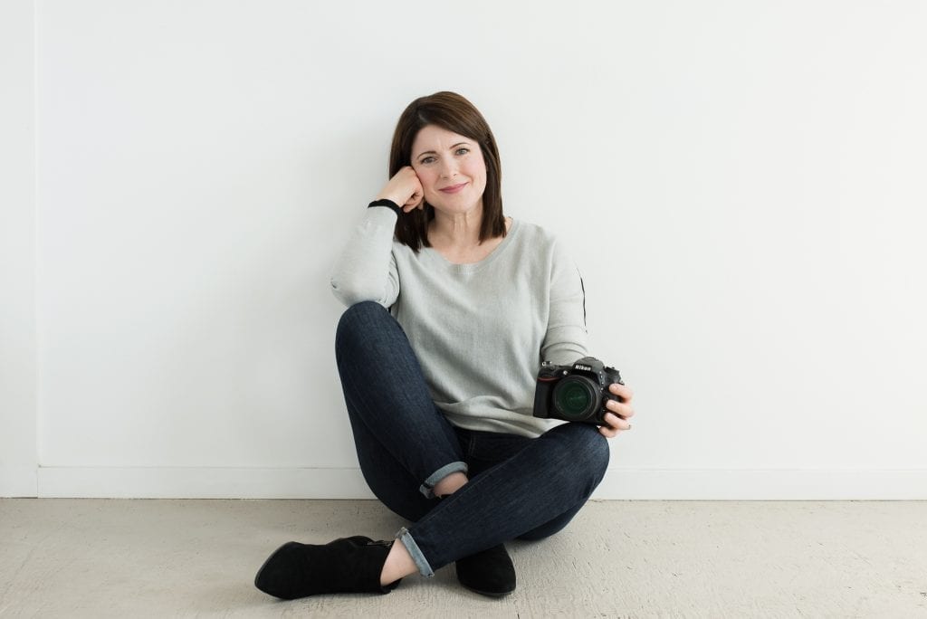 Rachelle sitting against a white wall with a camera in her hand